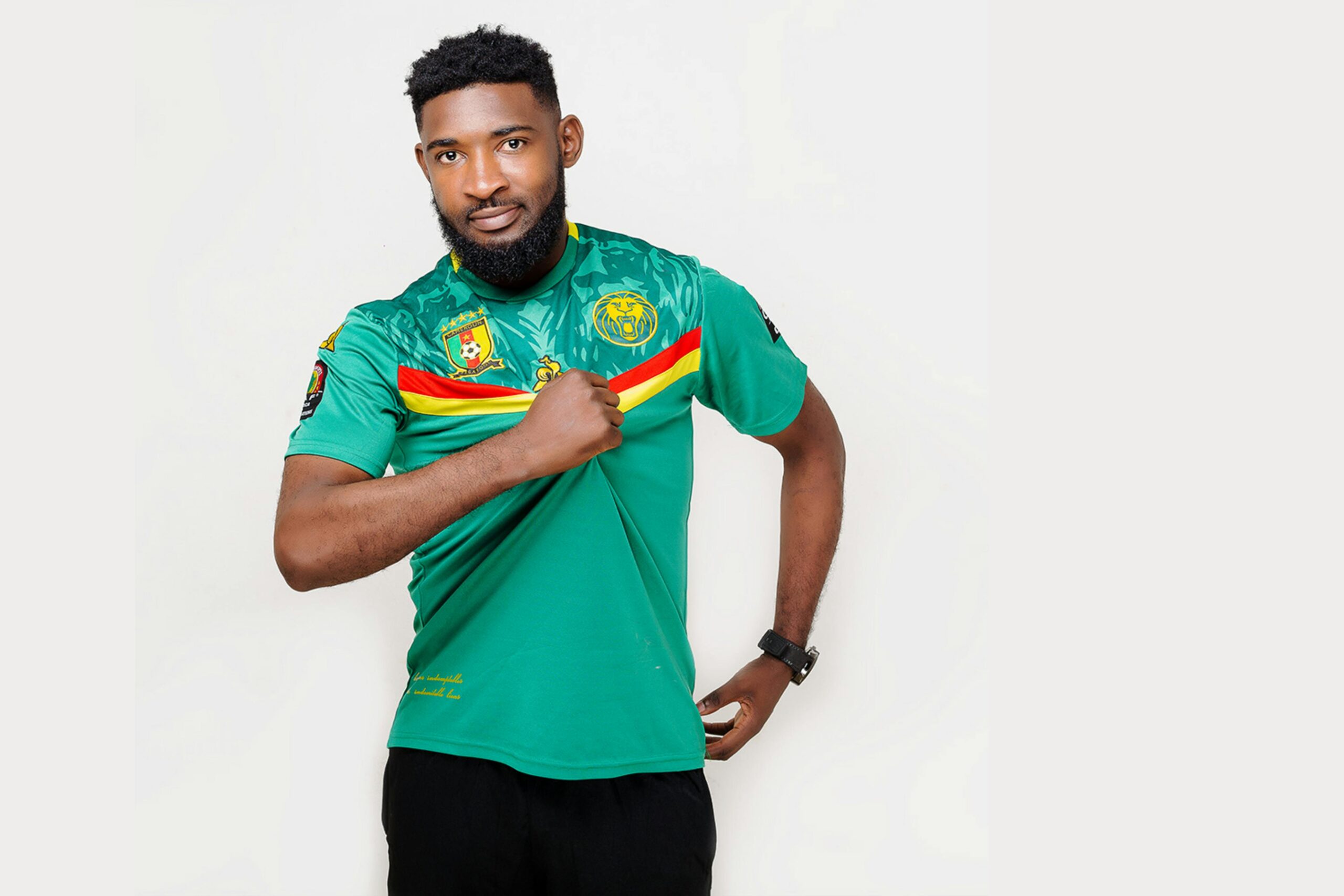 Confident African man in Cameroon football jersey with vibrant colors, posing against a white background.